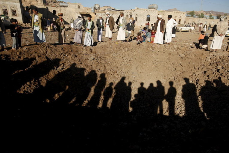 Люди стоят вокруг кратера после авиаудара на окраине столицы Йемена Саны