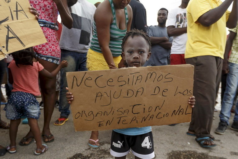 Африканский мальчик-мигрант на пограничном посту с Панамой держит плакат с надписью Нам нужна помощь от всемирной организации во время акции протеста в Пасо Каноас, Коста-Рика