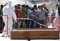 Мигранты стоят перед гробами после высадки с ирландского корабля ВМФ LE Niamh в сицилийском порту Мессина